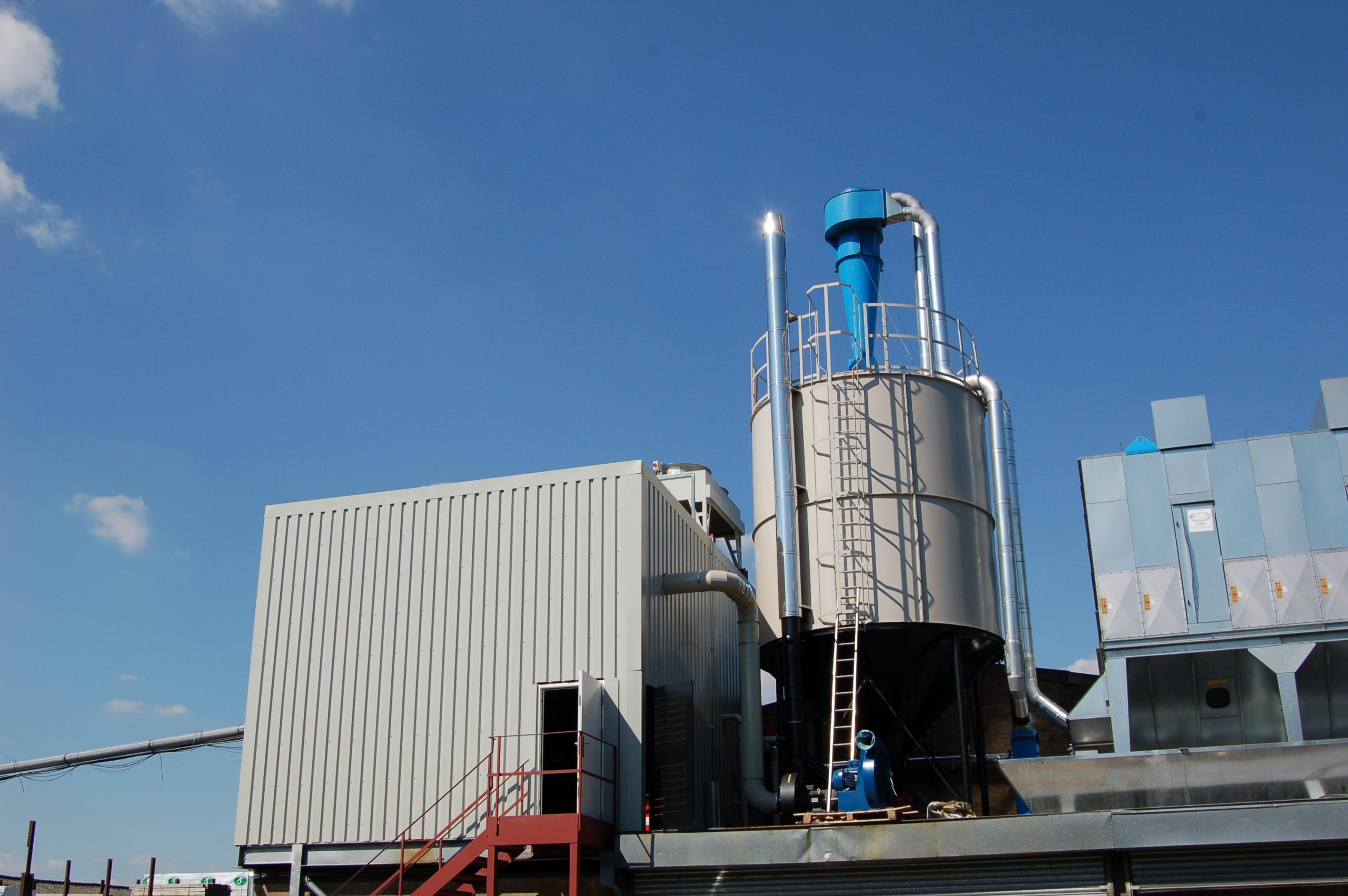 Site photograph of a Ranheat M15 biomass boiler in a boiler house, with mild steel conical silos and a chimney running up the side of the silo